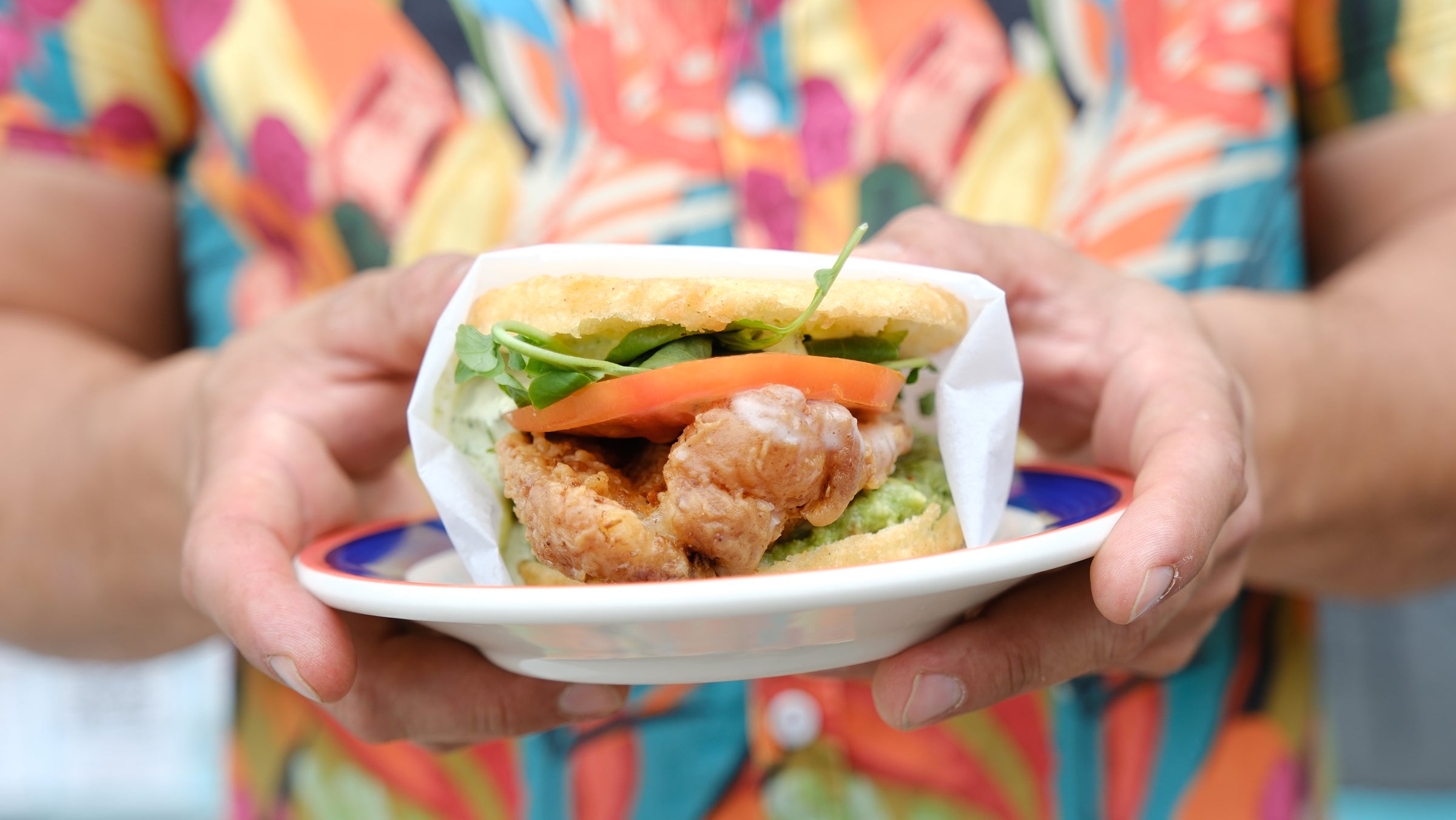 Crispy chicken arepa with watercress and tomato, held on a plate in a colorful tropical shirt at La Ñapa